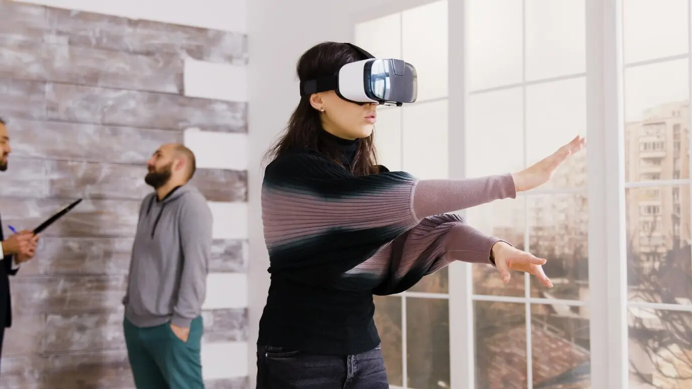 In einer leeren Wohnung nutzt eine Architektin eine Virtual-Reality-Brille, während im Hintergrund ein Immobilienmakler mit einem Kunden spricht.